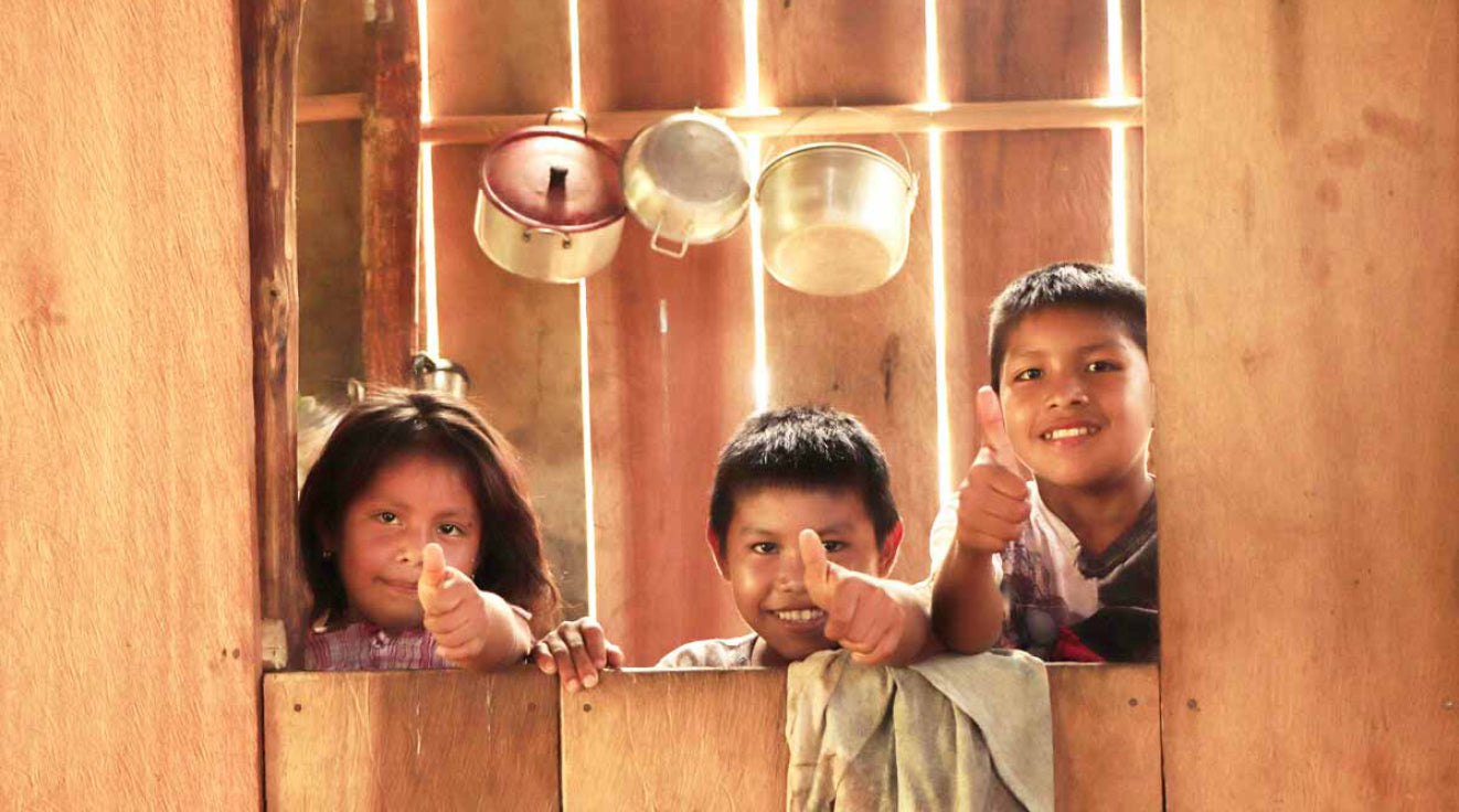 Drei Kinder die durch eine Öffnung in einer Holzwand schauen. Sie lächeln und zeigen Daumen hoch