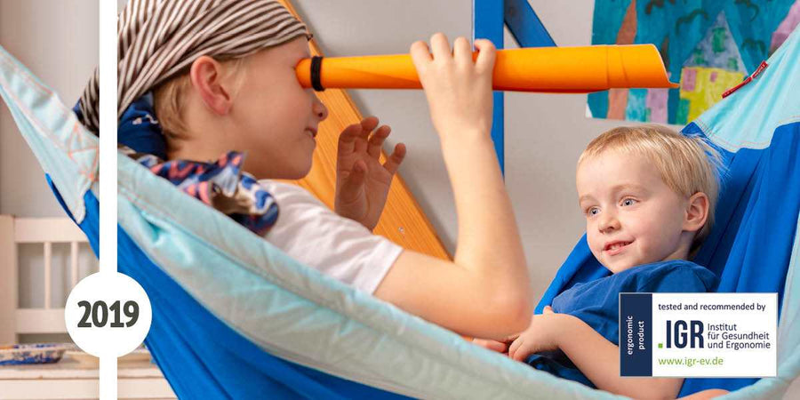 Zwei Kinder sitzen in einer blauen Kinderhängematte. Das eine hat ein Tuch um den Kopf gebunden und spiel mit einer Papierröhre Pirat